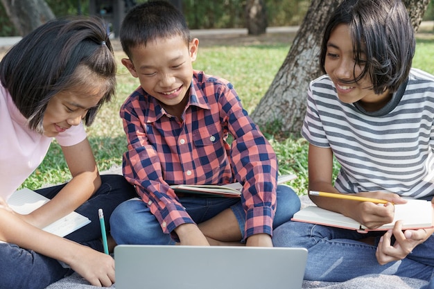 Grupo de niños de escuela primaria asiáticos que trabajan juntos y buscan información aprendiendo con laptop en el verano en el parque. La colegiala vino a estudiar viaje de campo y actividad aprendida al aire libre.