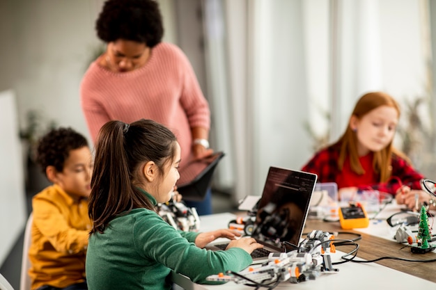 Grupo de niños felices con su profesora de ciencias afroamericana con programación de portátiles juguetes eléctricos y robots en el aula de robótica