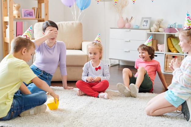 Grupo de niños pequeños alegres en gorras de cumpleaños sentados en círculo sobre una alfombra en la sala de estar mientras juegan con la mamá de uno de ellos