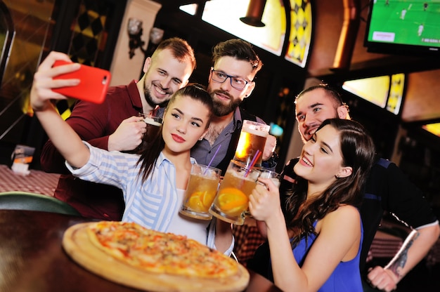 Grupo o compañía de jóvenes: los amigos beben cerveza, comen pizza, hablan y se ríen y se toman selfies en la cámara del teléfono inteligente en la superficie de la barra