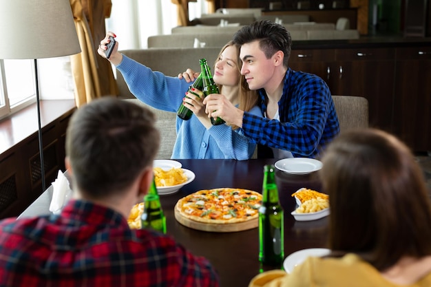 Grupo ou companhia de amigos jovens bebem cerveja comem pizza conversam e riem e tiram selfie