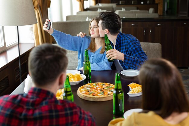 Grupo ou companhia de amigos jovens bebem cerveja comem pizza conversam e riem e tiram selfie