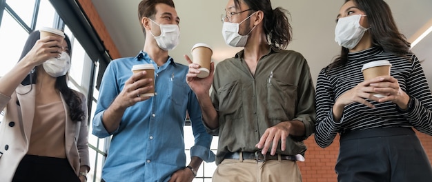 Grupo panorâmico de equipe de trabalhador de negócios com xícara de café para viagem, voltando ao escritório após a pausa para o almoço. Eles usam máscara protetora no novo consultório normal, evitando a propagação do coronavírus COVID-19.