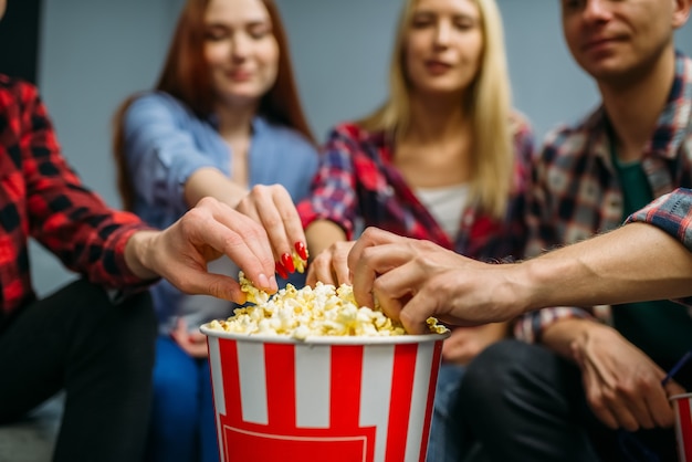 Grupo de personas comiendo palomitas de maíz y divirtiéndose en la sala de cine antes de la proyección. Jóvenes masculinos y femeninos sentados en el sofá en el cine