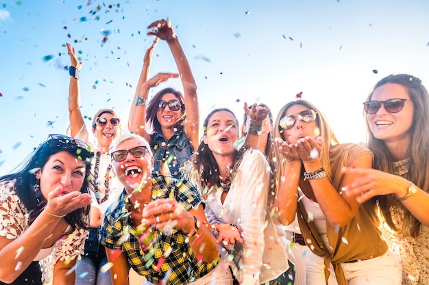 Grupo de personas felices, generaciones de edades mixtas, mujeres que se divierten todos juntos durante la fiesta de celebración - vista de demonios que soplan. confeti de colores y reír mucho en amistad