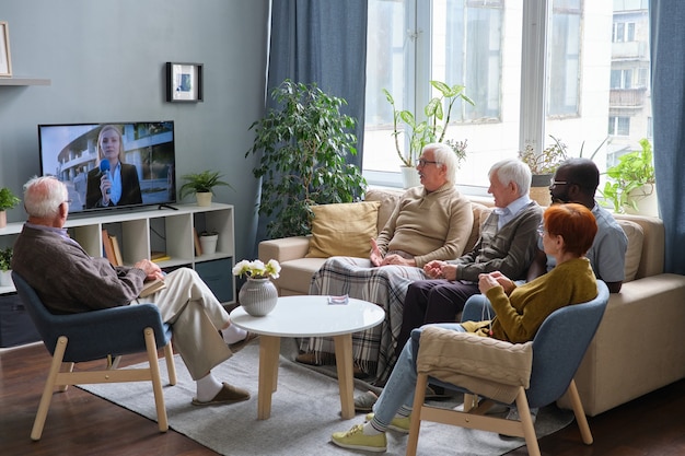 Grupo de personas mayores descansando en un sofá en la sala de estar y viendo las noticias en la televisión juntos