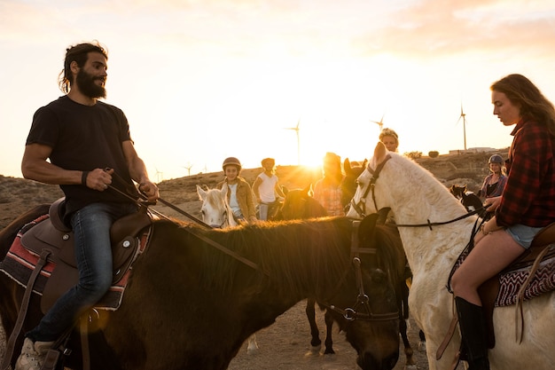 Grupo de personas montando caballos juntos en las colinas al atardecer - gente activa y feliz divirtiéndose y jugando con animales o caminando - vaquero y vaquera en un rancho