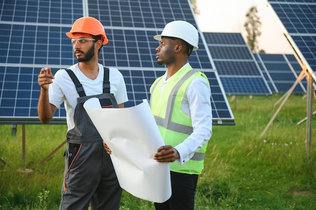 Grupo de personas multiétnicas con cascos de seguridad parados en la estación solar y mirando a la cámara sosteniendo grandes dibujos Dos ingenieros y técnicos examinando el plan de paneles al aire libre