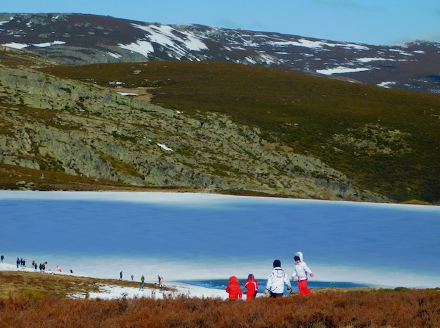 Un grupo de personas de pie frente a un lago con nieve en las montañas al fondo.