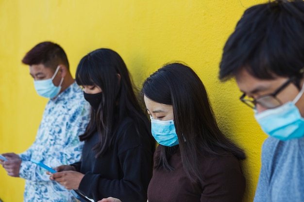 Grupo de personas que usan un teléfono móvil mientras usan una mascarilla durante la pandemia del virus corona frente a una pared amarilla