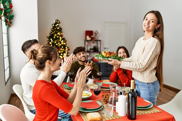 Grupo de personas reunidas aplaudiendo y sentadas en la mesa Mujer de pie y sosteniendo pavo asado celebrando la Navidad en casa