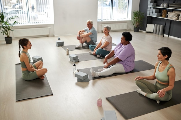 Grupo de personas sentadas en colchonetas de ejercicio y entrenando con instructor en clase de yoga