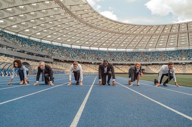 Grupo de personas vestidas con trajes de negocios de pie al comienzo de la pista deportiva preparándose para correr sprint