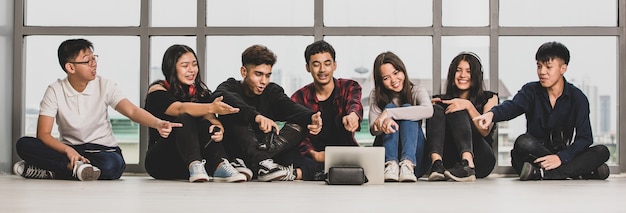 Grupo de siete adolescentes masculinos y femeninos en ropa casual sentados en el suelo y señalando con el dedo a la tableta. Estudiante universitario llamando por video en línea con amigos para entablar una conversación