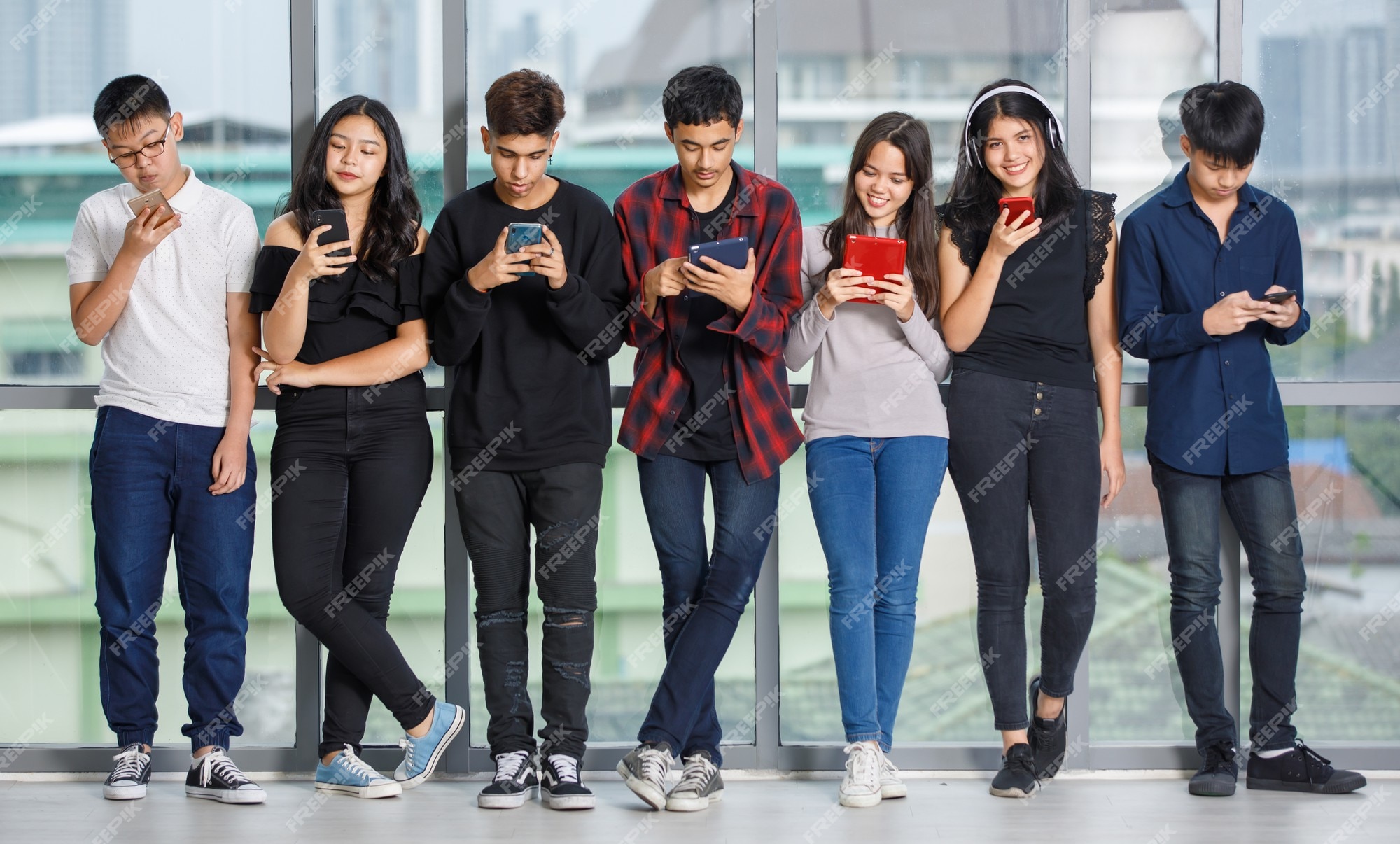 Grupo de siete adolescentes varones y mujeres jóvenes en ropa casual de pie y usando sus teléfonos inteligentes y tabletas. amigos la universidad jugando teléfonos móviles. concepto de teléfono adicto en