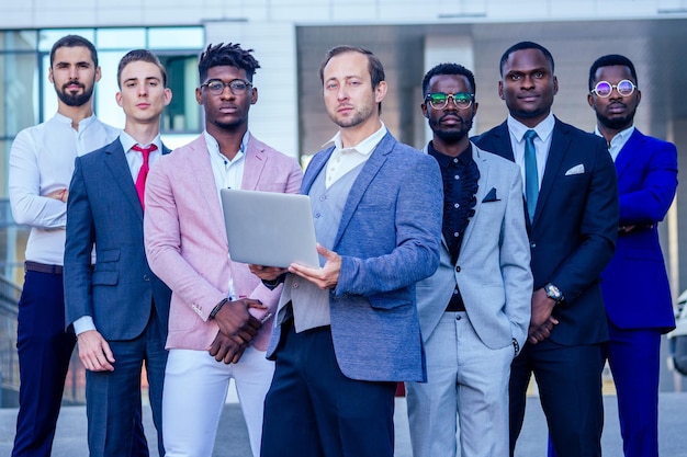 Un grupo de siete hombres de negocios exitosos con trajes elegantes de pie y mirando con orgullo al aire libre. trabajo en equipo y el concepto de empresa multiétnica