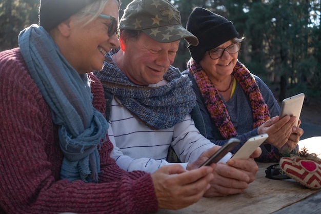 Grupo sonriente de personas sentadas al aire libre en el bosque en una mesa de madera usando su teléfono inteligente