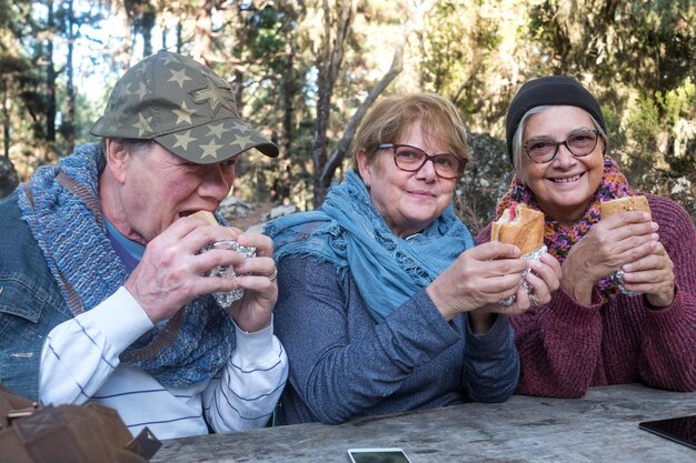 Grupo sonriente de tres amigos sentados al aire libre en el bosque comiendo sándwiches