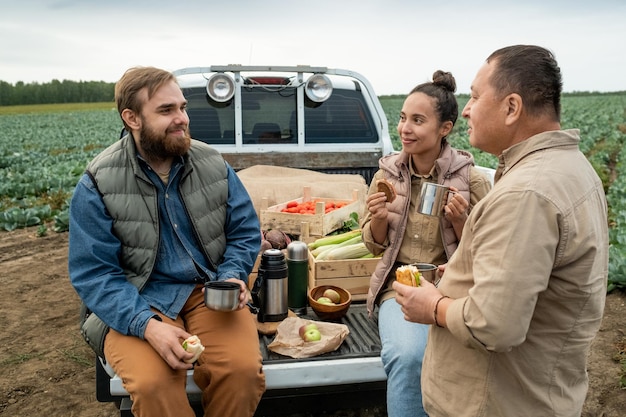 Grupo de trabajadores agrícolas multiétnicos sonrientes sentados en la cabina de una camioneta cerrada y bebiendo ...