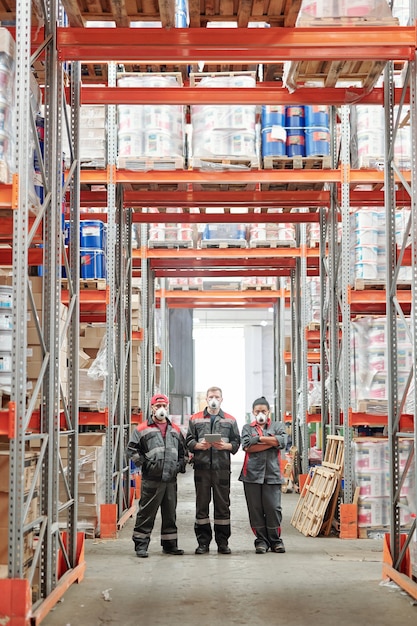 Grupo de trabajadores de grandes almacenes contemporáneos en guantes, respiradores y ropa de trabajo de pie en el pasillo entre bastidores con cajas llenas de mercancías