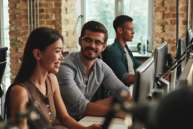 Grupo de trabajo en equipo multiétnico de empleados jóvenes alegres que trabajan en computadoras y hablan con cada uno