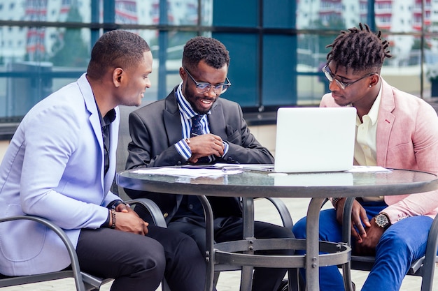 Un grupo de tres elegantes empresarios afroamericanos socios empresarios en trajes de negocios de moda reunidos sentados a la mesa firman valores con un portátil en un café de verano al aire libre