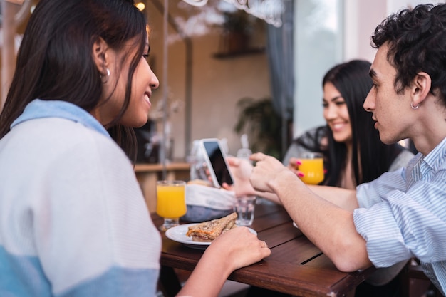 Grupo de tres estudiantes latinos sentados en la barra de una cafetería mirando un teléfono inteligente.