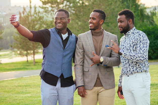 Un grupo de tres hombres negros con elegantes trajes en un parque de verano. Amigos afroamericanos empresario hispano se fotografiaron selfie en el teléfono al aire libre