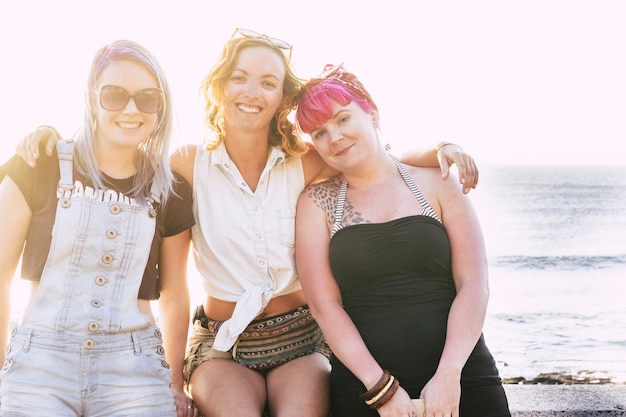grupo de tres mujeres abrazados y sentados en la playa mirando y sonriendo con el mar y el agua azul