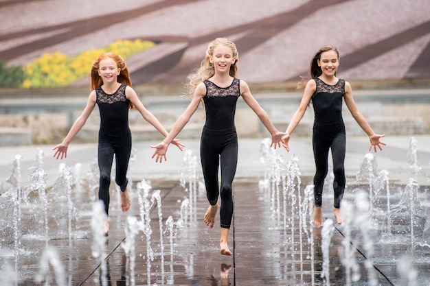 Un grupo de tres pequeñas bailarinas con trajes negros ajustados corren hacia el espectador en medio de fuentes salpicando contra el fondo de un paisaje urbano en un día caluroso.