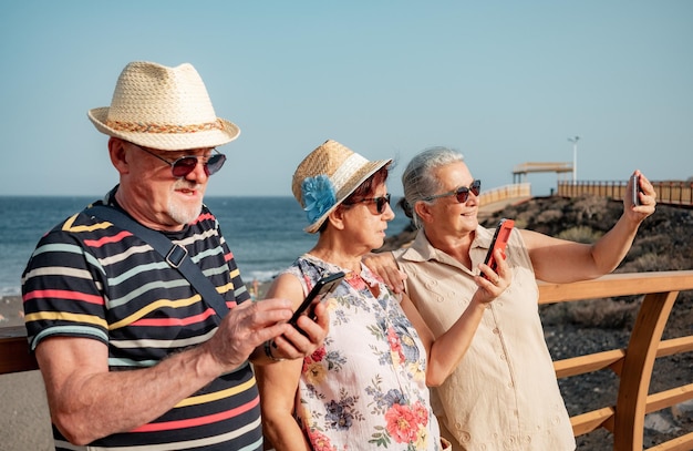 Grupo de tres personas mayores con sombreros y gafas de sol usando un teléfono móvil al aire libre en el mar Felices jubilados disfrutando del tiempo libre en un día soleado