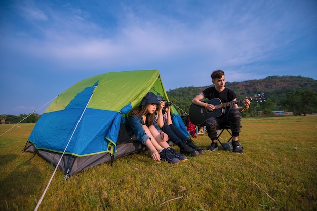 Grupo de turistas asiáticos amigos bebiendo y tocando la guitarra junto con la felicidad en verano mientras acampa cerca del lago al atardecer