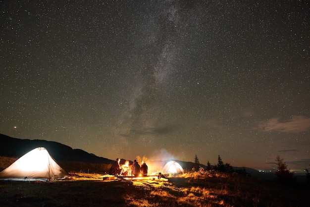 Grupo de turistas con guitarra quemando hoguera bajo el oscuro cielo estrellado con la constelación de la Vía Láctea.