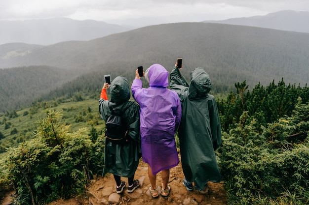 Grupo de viajeros en impermeables en la montaña toma fotos de la naturaleza en los teléfonos.