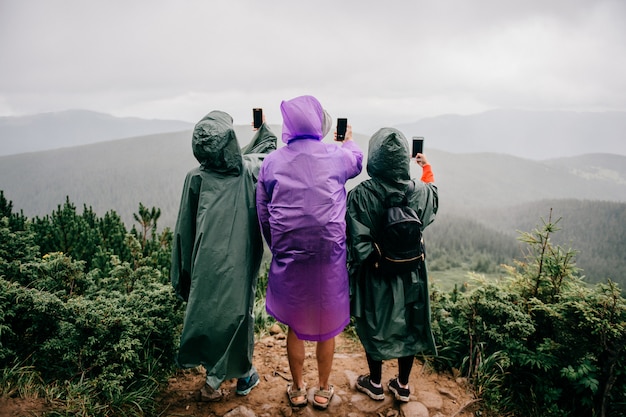 Grupo de viajeros en impermeables se paran en las montañas y toman fotos del paisaje de la naturaleza salvaje en los teléfonos. Tres turistas hacen seilfie con teléfono en día lluvioso de verano