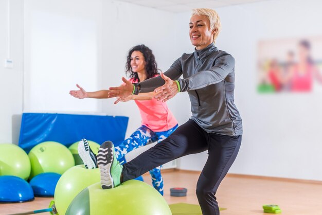 Gruppe aktiver, fröhlicher, sportlicher Frauen, die im Fitnessstudio einbeinige Kniebeugen mit Balance-Ball-Training machen