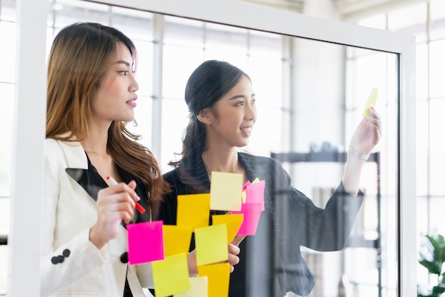 Gruppe erfolgreicher Teamwork asiatischer Geschäftsfrauen Brainstorming-Meeting mit bunten Haftnotizen an der Glaswand für neue Ideen mit agiler Methodik für Unternehmen in einem Tech-Startup-Büro