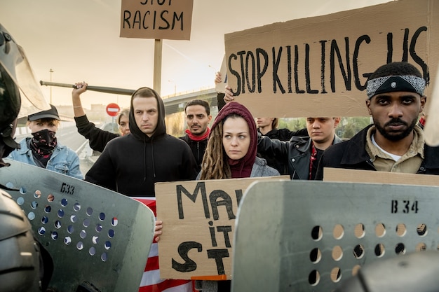 Gruppe ernsthafter junger multiethnischer Aktivisten, die mit Schildern auf der Straße stehen und zusammen protestieren, Polizeiwachen, die sie mit Schilden stoppen