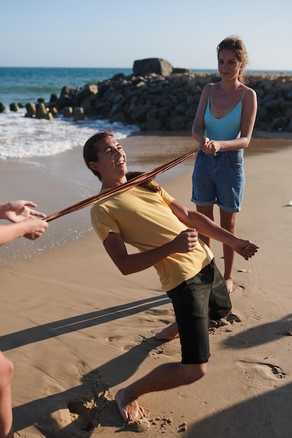 Gruppe fröhlicher Freunde, die ein Limbo-Spiel am Strand-Sommerurlaubskonzept spielen