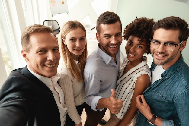 Gruppe fröhlicher Kollegen, die Selfies machen und gestikulieren, während sie im modernen Büro stehen