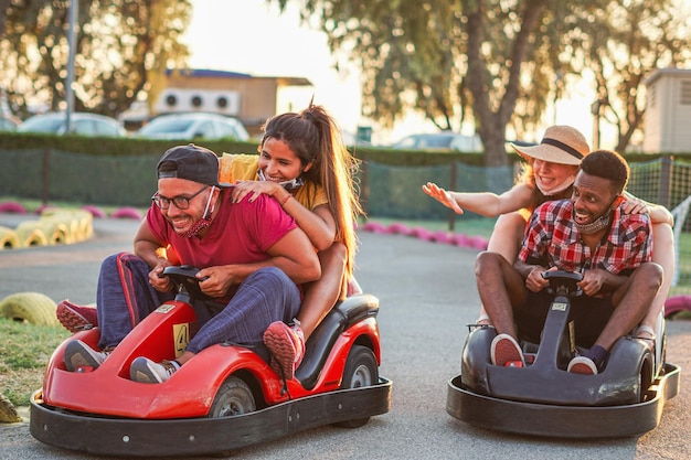 Gruppe gemischtrassiger Freunde, die sich mit Go-Kart amüsieren Junge Leute mit Gesichtsmaske lächelnd und fröhlich bei Mini-Autorennen Paare draußen bei Doppeldate Neuer Lebensstil