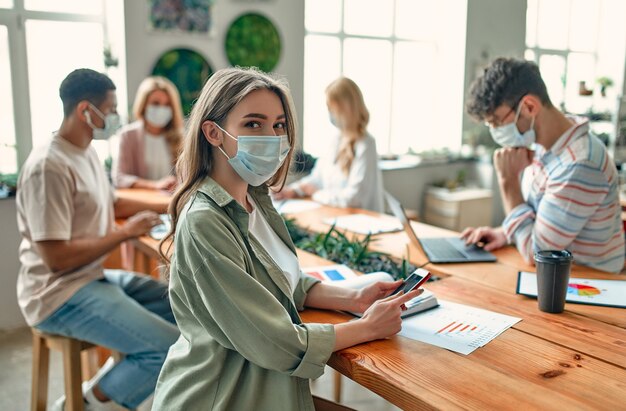 Gruppe gemischtrassiger Menschen, die während des Coronavirus in einem modernen Büro arbeiten. Junge Geschäftsleute und Senior-Chef arbeiten zusammen und tragen Gesichtsschutzmasken. Vorbeugende Maßnahmen während Covid-19