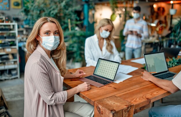 Gruppe gemischtrassiger Menschen, die während des Coronavirus in einem modernen Büro arbeiten. Junge Geschäftsleute und Senior-Chef arbeiten zusammen und tragen Gesichtsschutzmasken. Vorbeugende Maßnahmen während Covid-19