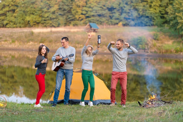 Gruppe glückliche Freunde mit Gitarre, Spaß im Freien habend, tanzen und springen nahe dem See im Park