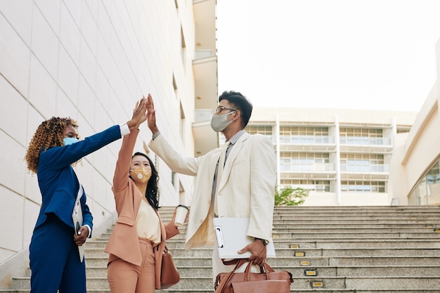 Gruppe glücklicher aufgeregter Geschäftsleute in medizinischen Masken, die im Freien stehen und sich nach Abschluss der Arbeit an einem großen Projekt gegenseitig High Five geben