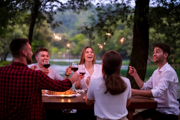 gruppe glücklicher freunde, die ferien mit sprinklern feiern und rotwein trinken, während sie an einem schönen sommerabend in der natur ein französisches picknick-abendessen im freien in der nähe des flusses haben