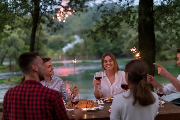 gruppe glücklicher freunde, die rotweinglas toasten, während sie während der sommerferienferien in der nähe des flusses in der wunderschönen natur ein picknick-französisches abendessen im freien haben