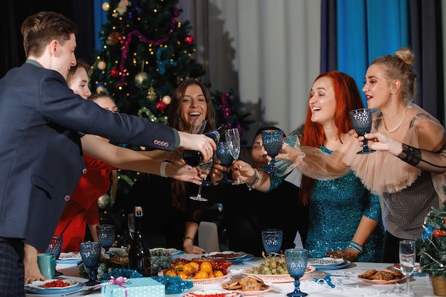 Gruppe glücklicher Freunde, die Weihnachten oder Neujahr feiern. Ein Mann gießt Champagner in Gläser. Frauen lachen, sitzen an einem Tisch neben dem Weihnachtsbaum.