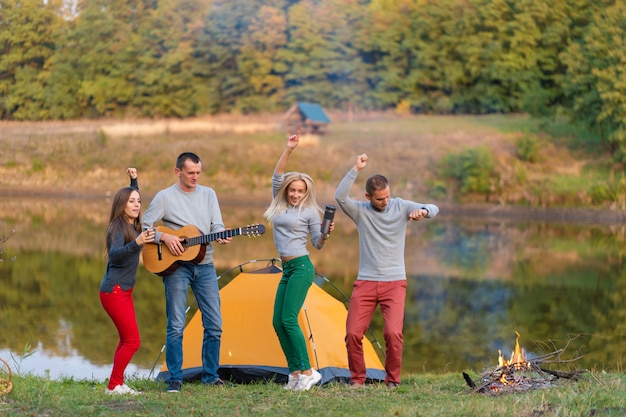 Gruppe glücklicher Freunde mit Gitarre, die Spaß im Freien haben, tanzen und in der Nähe des Sees im Parkhintergrund den schönen Himmel springen. Camping Spaß