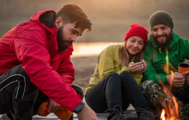 Gruppe glücklicher junger wanderer in warmer aktivkleidung, die nahe lagerfeuer sitzen und heißes getränk trinken, während sie sich nach dem trekking in der natur am herbstabend ausruhen